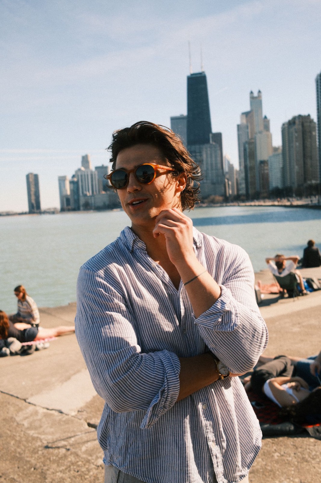 Logan in a striped linen shirt and tortoiseshell sunglasses on the Chicago lakefront, with the skyline behind him